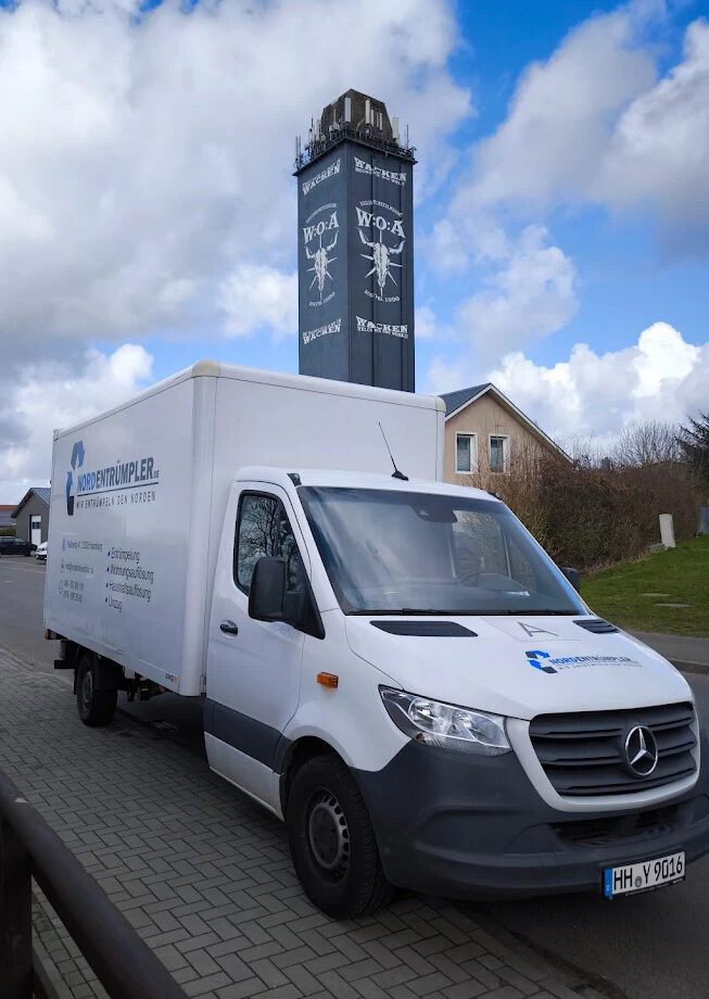 Ein weißer Mercedes-Benz-Lieferwagen parkt auf einer mit Ziegeln gepflasterten Straße. Dahinter steht ein hoher, dunkler Turm mit weißer Bemalung und der Aufschrift "W/O-A". Der Himmel ist teilweise bewölkt, und im Hintergrund sind Häuser mit roten Dächern zu sehen.