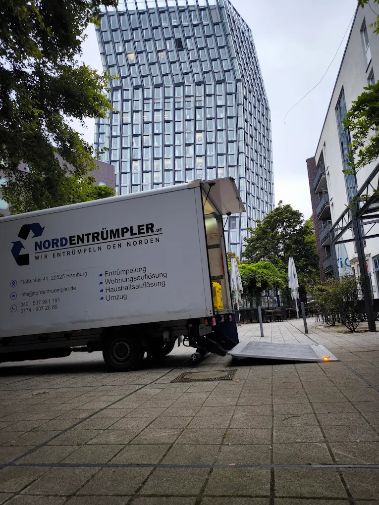 Ein weißer Umzugswagen mit dem Schriftzug "NORDENTRÜMPLER" ist auf einem gepflasterten Platz neben hohen Gebäuden und Bäumen geparkt. Seine hintere Rampe ist nach unten ausgefahren, die Rückseite ist offen. Ein modernes gläsernes Hochhaus dominiert den Hintergrund unter einem bewölkten Himmel.