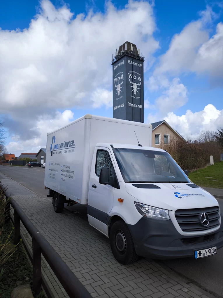 Ein weißer Mercedes-Benz Lieferwagen ist auf einer gepflasterten Straße neben einem Geländer geparkt. Im Hintergrund ist ein hoher schwarzer Turm mit weißer Grafik und Text zu sehen. Einige Häuser und Bäume sind unter einem teilweise bewölkten Himmel zu sehen.