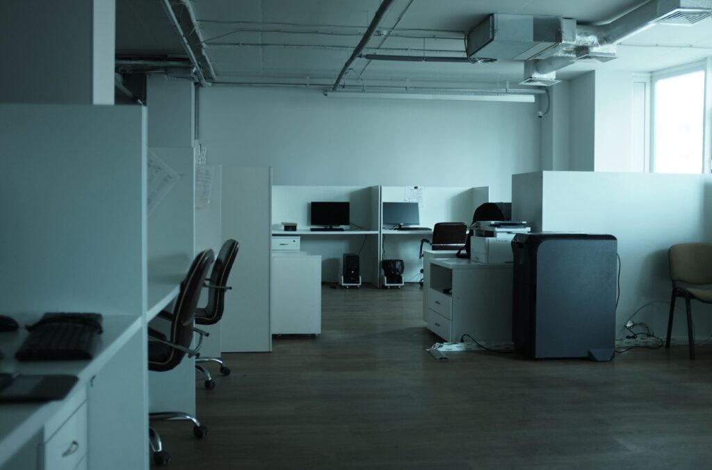 In einem schwach beleuchteten, leeren Büro stehen mehrere Arbeitsnischen mit Computern, Schreibtischen, Stühlen und Bürogeräten wie einem Kopierer. Der Boden besteht aus Holz, und Tageslicht fällt durch ein großes Fenster auf der rechten Seite des Bildes herein.