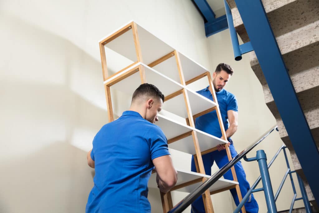 Zwei Umzugshelfer in blauen Uniformen tragen ein hohes Bücherregal aus weißem Holz eine Treppe hinauf. Man sieht sie von unten, wie sie das Holzmöbelstück vorsichtig die Betontreppe mit Metallgeländer hinauftragen.