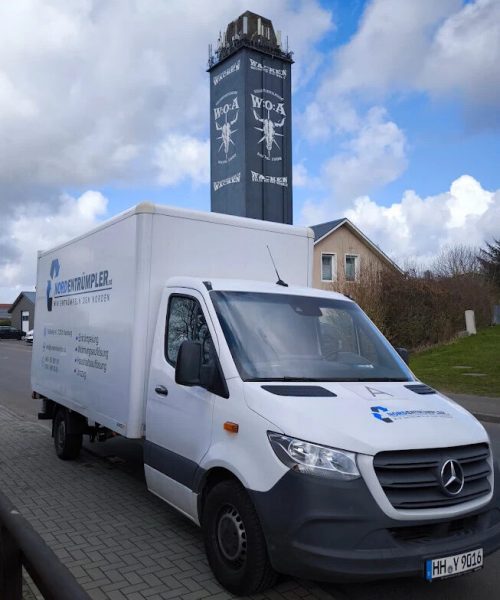 Ein weißer Mercedes-Benz-Lieferwagen parkt auf einer mit Ziegeln gepflasterten Straße. Dahinter steht ein hoher, dunkler Turm mit weißer Bemalung und der Aufschrift "W/O-A". Der Himmel ist teilweise bewölkt, und im Hintergrund sind Häuser mit roten Dächern zu sehen.