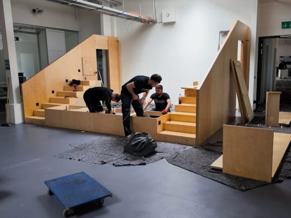 Drei Personen sind dabei, in einem modernen Industrieraum mit grauen Böden, freiliegenden Rohren und Oberlichtern eine Holzkonstruktion zusammenzubauen, die an Treppen und Plattformen erinnert. Werkzeuge und Materialien liegen in der Nähe verstreut, darunter eine Plattform auf Rädern und eine große schwarze Tasche.