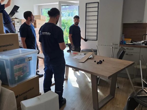 Vier Umzugshelfer sind in einem sonnendurchfluteten Raum dabei, Möbel zu montieren oder zu demontieren. Einer hält ein Metallbettgestell, die anderen stehen an einem unordentlichen Tisch. Auf dem Holzboden sind Kartons, Stühle und verstreute Werkzeuge zu sehen, und eine offene Terrassentür lässt das Tageslicht herein.