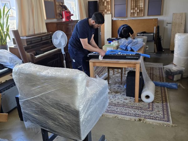 Zwei Personen wickeln auf einem Holztisch in einem Raum, in dem sich ein eingepackter Sessel, ein Klavier und Verpackungsmaterial befinden, eine Tastatur in Luftpolsterfolie ein. Sonnenlicht strömt durch die Fenster; ein Teppich bedeckt einen Teil des Bodens. Der Raum scheint ein Atelier oder ein Arbeitsraum zu sein.