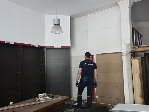 Ein Mann in dunkelblauer Arbeitskleidung steht in einem unordentlichen Raum, in dem Holzstücke und Trümmer verstreut liegen. Er steht mit dem Rücken zur Kamera und blickt auf eine kahle Wand. Über ihm hängen zwei Deckenlampen. Im Vordergrund steht ein großer Holztisch, der teilweise von Trümmern bedeckt ist.