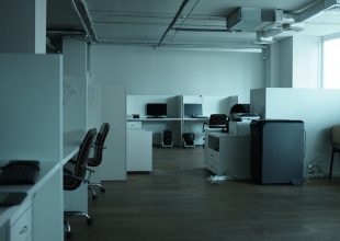In einem schwach beleuchteten, leeren Büro stehen mehrere Arbeitsnischen mit Computern, Schreibtischen, Stühlen und Bürogeräten wie einem Kopierer. Der Boden besteht aus Holz, und Tageslicht fällt durch ein großes Fenster auf der rechten Seite des Bildes herein.
