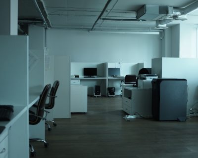 In einem schwach beleuchteten, leeren Büro stehen mehrere Arbeitsnischen mit Computern, Schreibtischen, Stühlen und Bürogeräten wie einem Kopierer. Der Boden besteht aus Holz, und Tageslicht fällt durch ein großes Fenster auf der rechten Seite des Bildes herein.