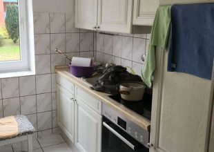 Die Küche verfügt über cremefarbene Schränke, eine geflieste Küchenrückwand und ein Fenster mit Blick auf den Rasen. Auf der Arbeitsplatte stehen ein violettes Waschbecken, ein Topf und ein Stapel Stoff, während neben dem Fenster ein Hocker steht und ein blaues Handtuch am Schrank hängt.