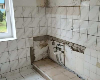 Eine teilweise geflieste Küchenecke mit freiliegenden Wasserleitungen, fehlenden Schränken und Geräten. An der Wand sind Reste von altem Kleber und Flecken zu sehen. Der Boden ist weiß gefliest, und links befindet sich ein Fenster, durch das Tageslicht hereinfällt.
