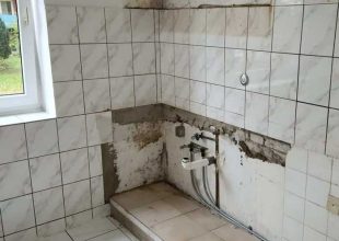 Eine Küche, die gerade renoviert wird, mit weiß gefliesten Wänden und Boden. Ein Teil der gefliesten Wand wurde entfernt, wodurch roher Putz und freiliegende Wasserleitungen in der Nähe einer kleinen, erhöhten, gefliesten Plattform zum Vorschein kommen.