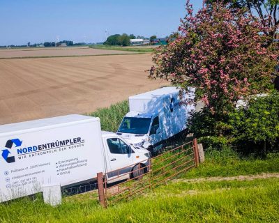 Drei weiße Service-Transporter mit dem „Nordentrümpler“-Logo stehen auf einem ländlichen Feldweg neben einem blühenden Baum und einem Haus, im Hintergrund erstrecken sich weite Felder vor einem blauen Himmel. Die Szene wirkt sonnig und friedlich.