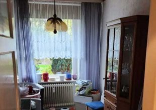 Ein Fenster mit grauen Vorhängen bietet einen Blick auf einen Hof. Darunter befinden sich ein Heizkörper und mehrere Topfpflanzen, rechts davon steht ein hoher Geschirrschrank aus Holz.